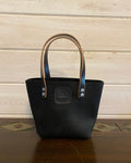 A black leather tote bag with tan handles and silver-tone hardware, standing on a dark wooden surface against a light wood plank wall.