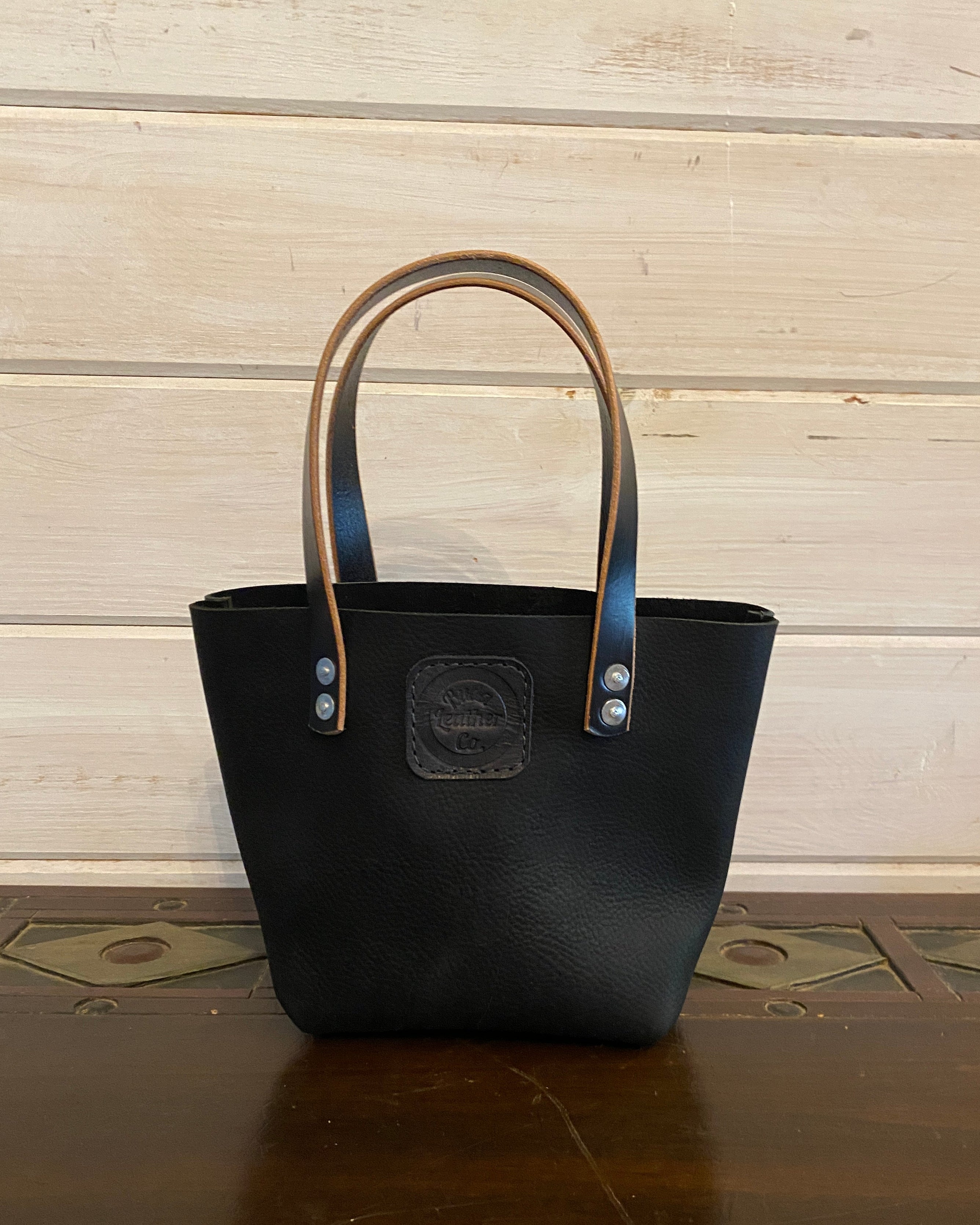 A black leather tote bag with tan handles and silver-tone hardware, standing on a dark wooden surface against a light wood plank wall.