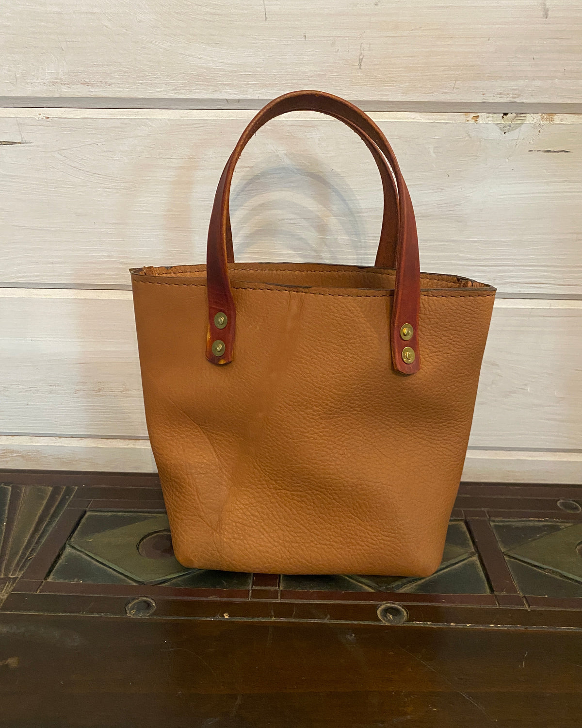 A tan pebbled leather tote bag with reddish-brown leather handles and brass-tone rivets, photographed standing upright against a white wood-paneled wall next to a dark wooden surface.