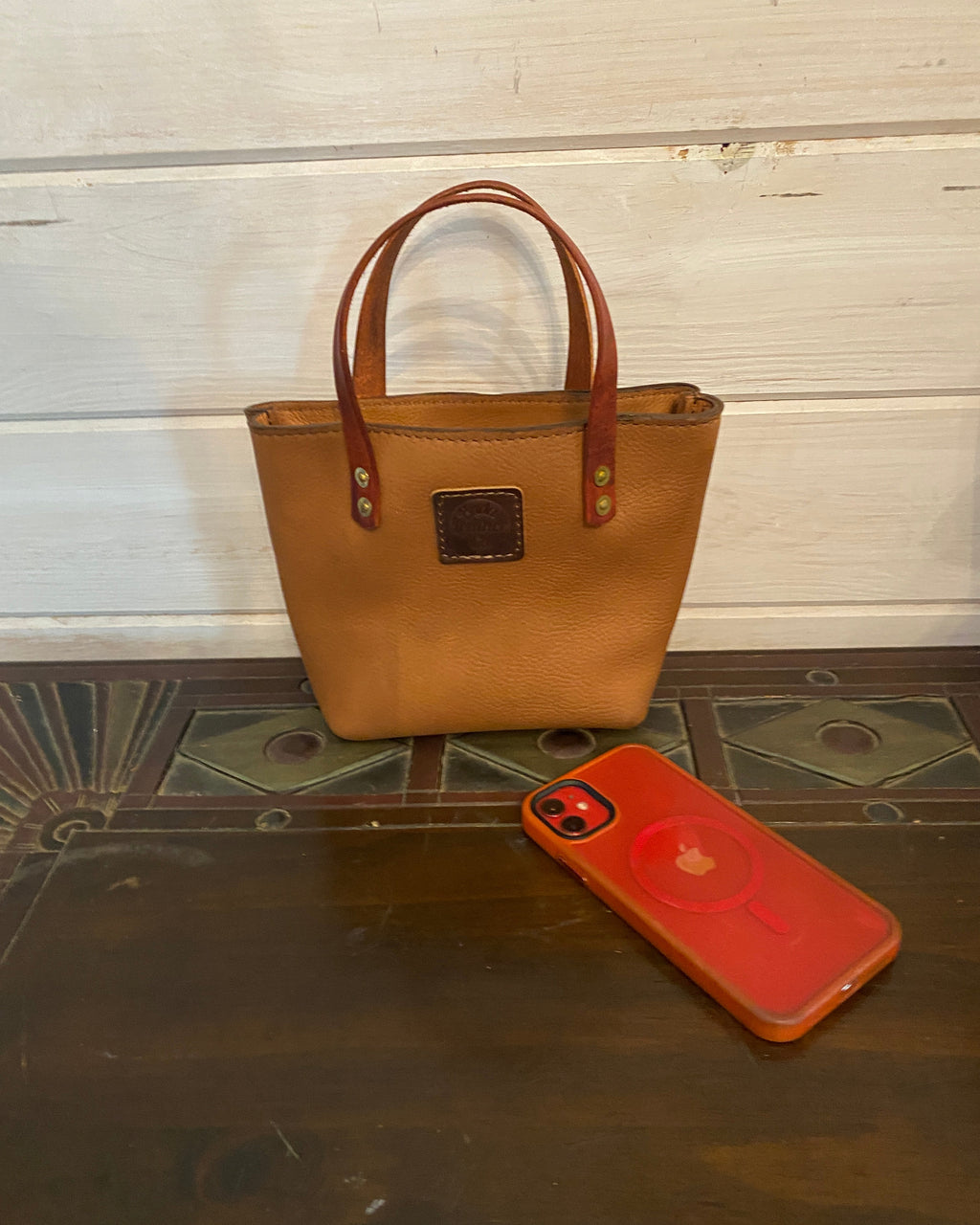 A tan leather tote bag with brown leather handles, shown next to a red phone case on a dark wood floor and white wooden panel wall.