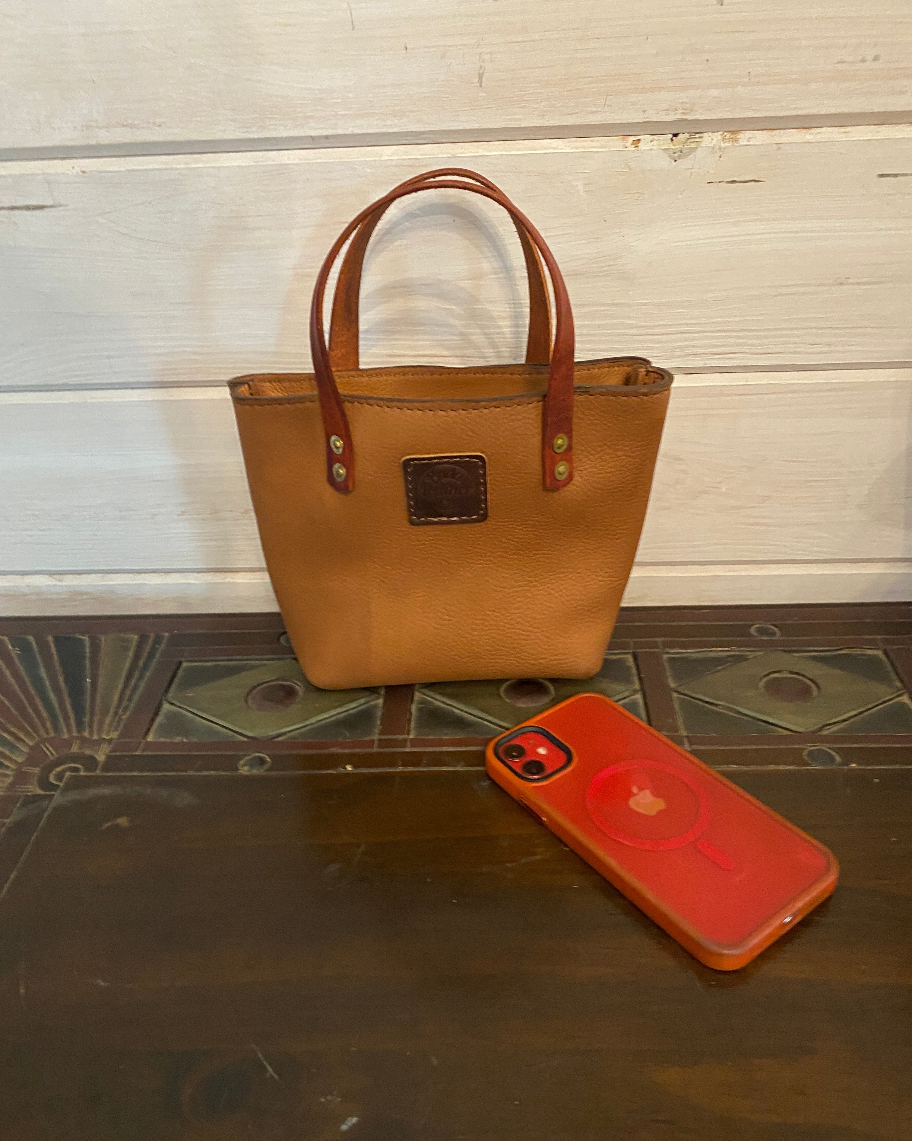 A tan leather tote bag with brown leather handles, shown next to a red phone case on a dark wood floor and white wooden panel wall.