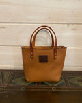 A tan leather tote bag with dark brown handles and a brown logo patch on the front, standing on a dark tiled surface against a light wood paneled wall.