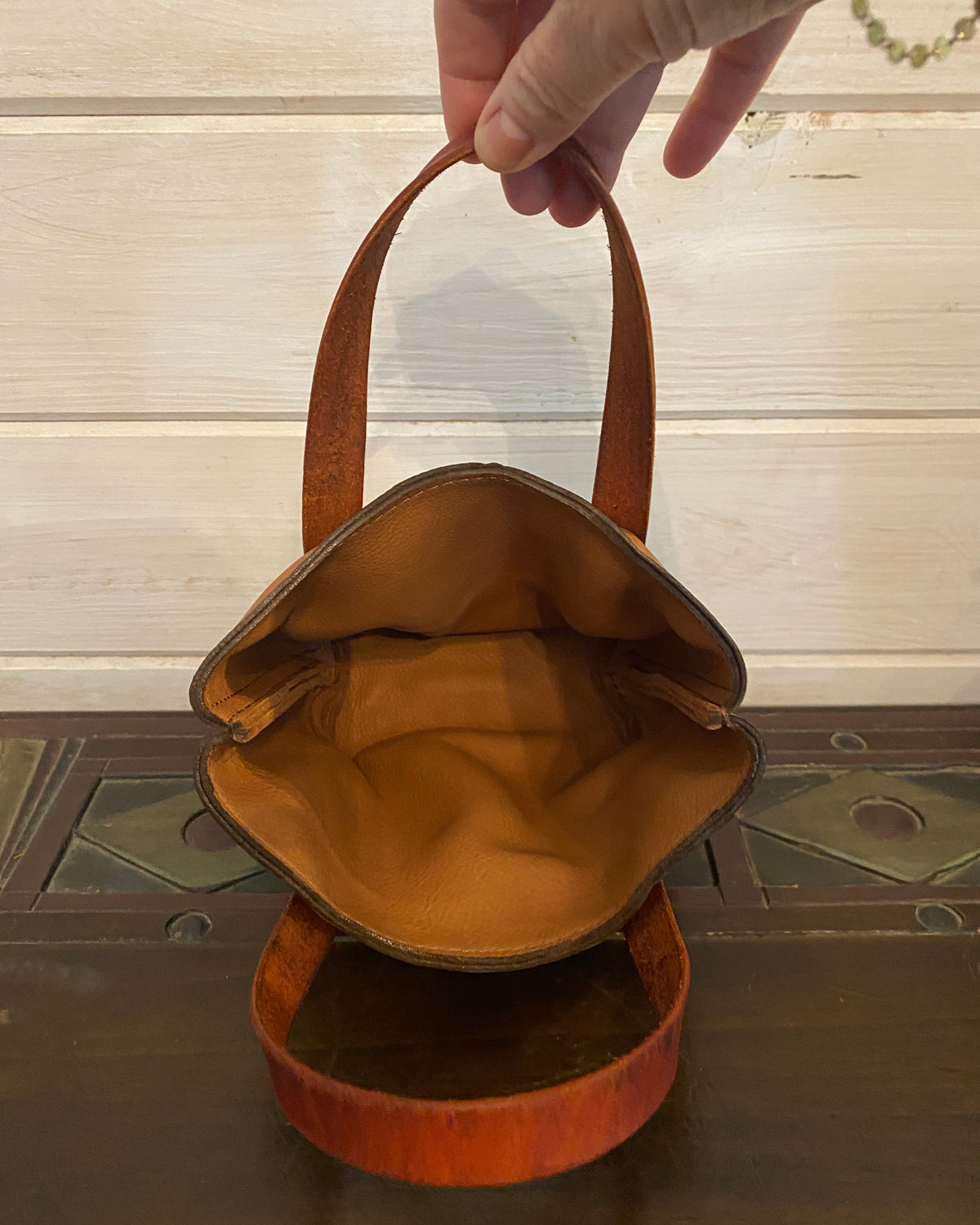 A hand holding the handle of a small brown leather shoulder bag with a tan interior lining, shown against a light wood paneled wall and dark wooden furniture.