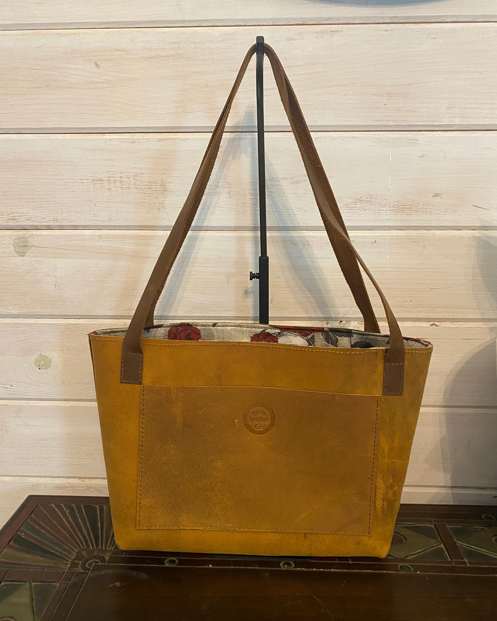 A tan leather tote bag with a floral patterned lining and brown shoulder straps, shown from a side angle against a white wood paneled wall.