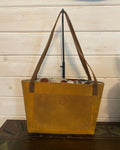 A tan leather tote bag with a floral patterned lining and brown shoulder straps, shown from a side angle against a white wood paneled wall.