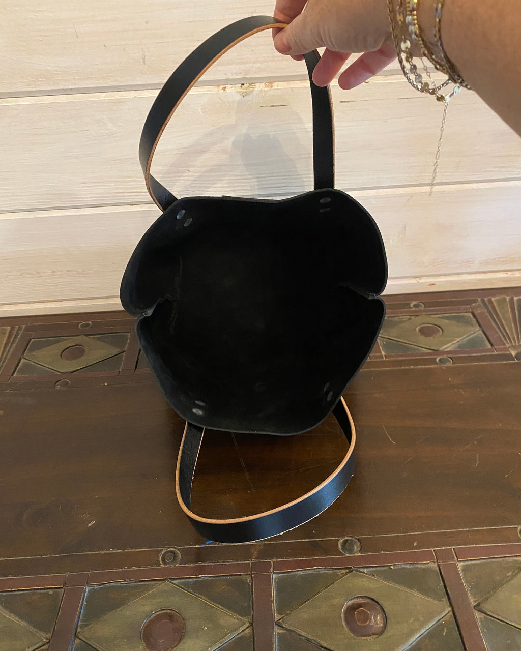 A black tote with a curved top handle, held open by a person's hand. The bag is resting on a dark wooden surface with a light wood paneled wall in the background.