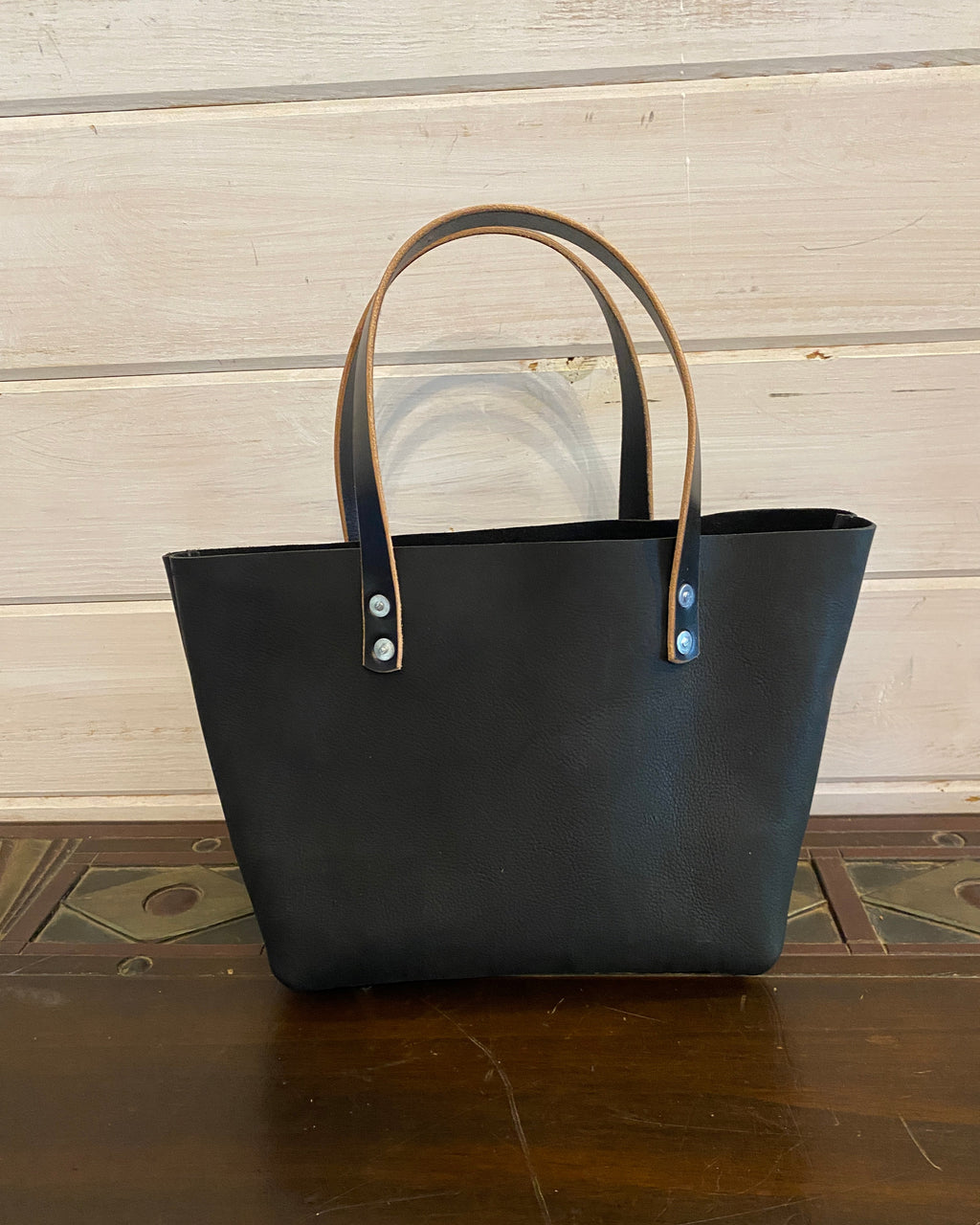 A black leather tote bag with tan leather handles and silver-tone rivets, standing on a dark wooden surface against a light wood paneled wall.