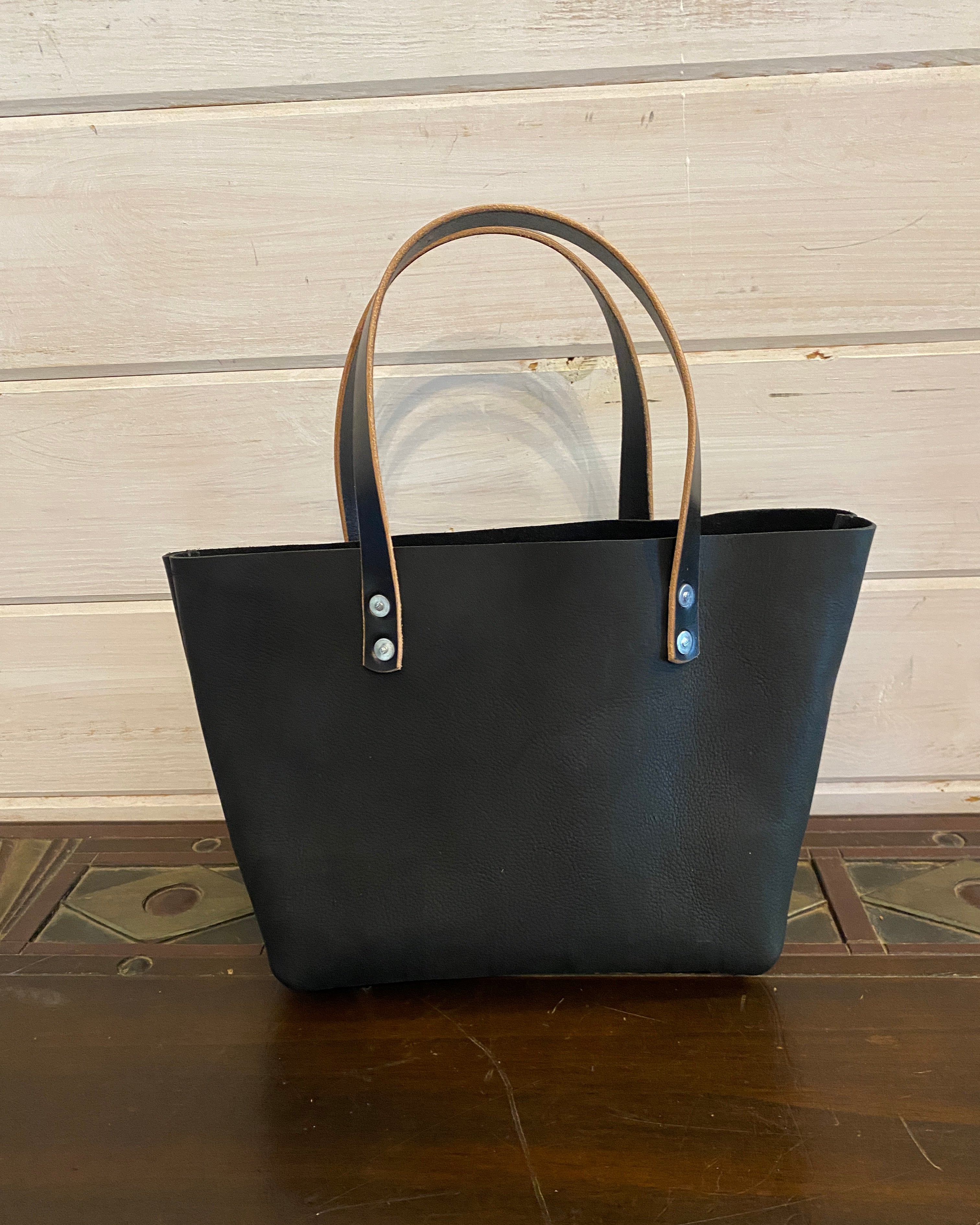 A black leather tote bag with tan leather handles and silver-tone rivets, standing on a dark wooden surface against a light wood paneled wall.