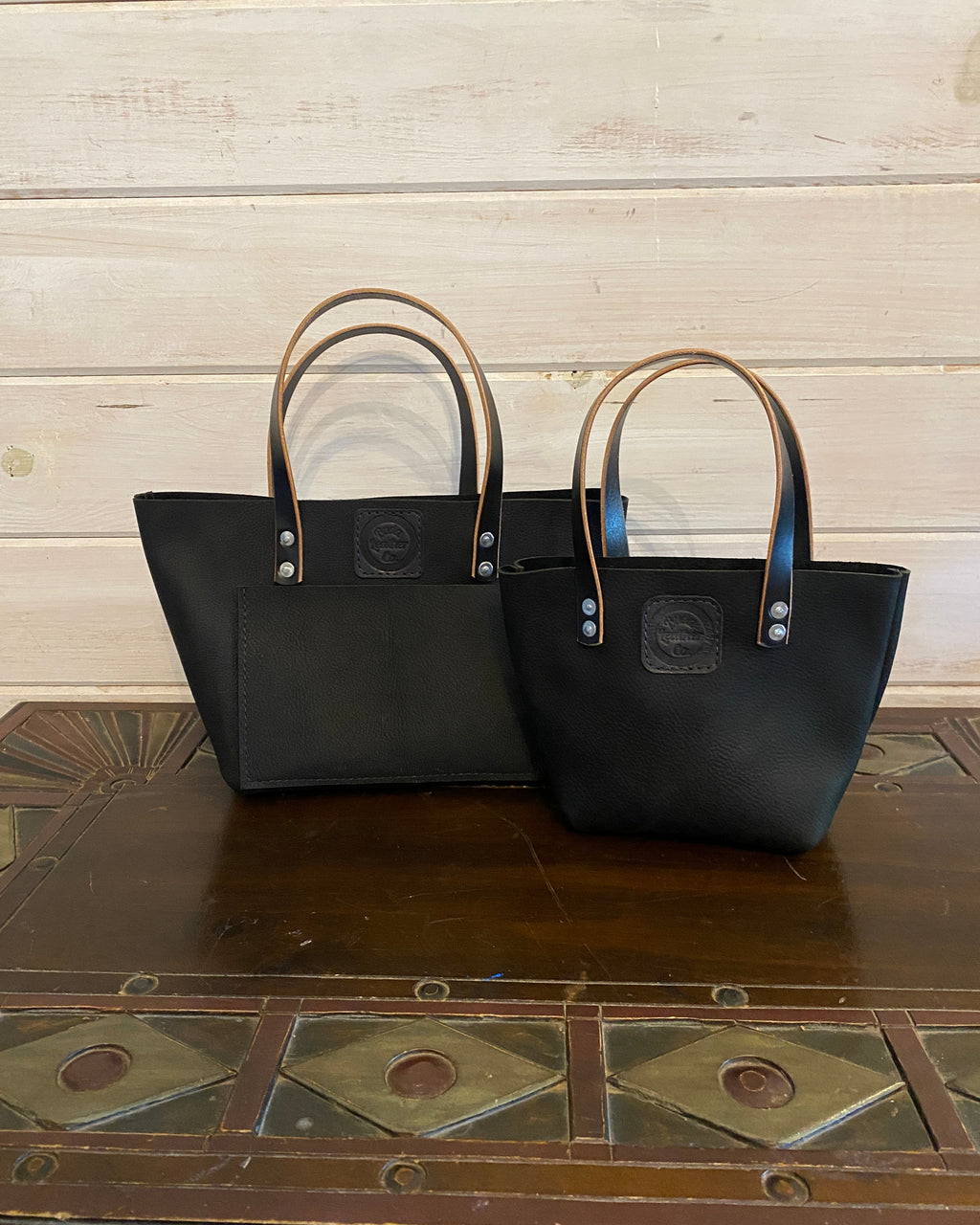 Two black leather tote bags with tan handles, displayed side by side on a rustic wooden table against a light wood plank wall.