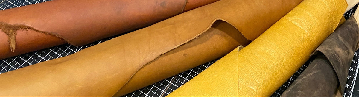 A leatherworking workshop with multiple rolls of leather in various colors, including tan, brown, yellow, and light blue, hanging on wooden racks and laid out on a workbench.
