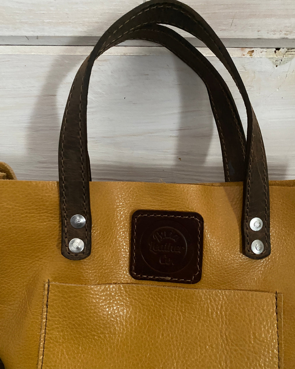 Close-up of a tan pebbled leather tote bag with dark brown leather handles and a matching dark brown square logo patch on a white wooden surface.