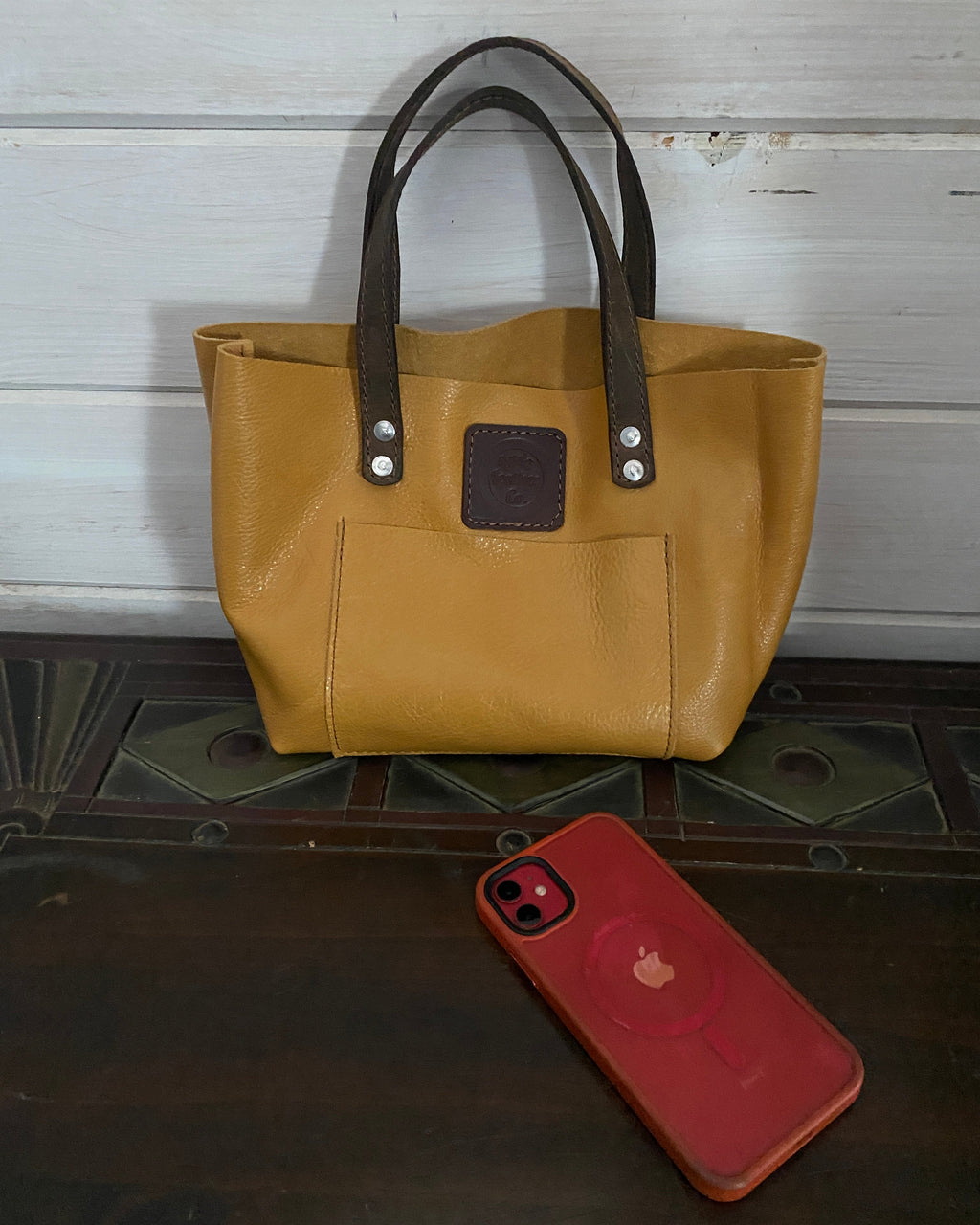A tan leather tote bag with dark brown handles and a front pocket, shown next to a red smartphone on a dark wooden surface.
