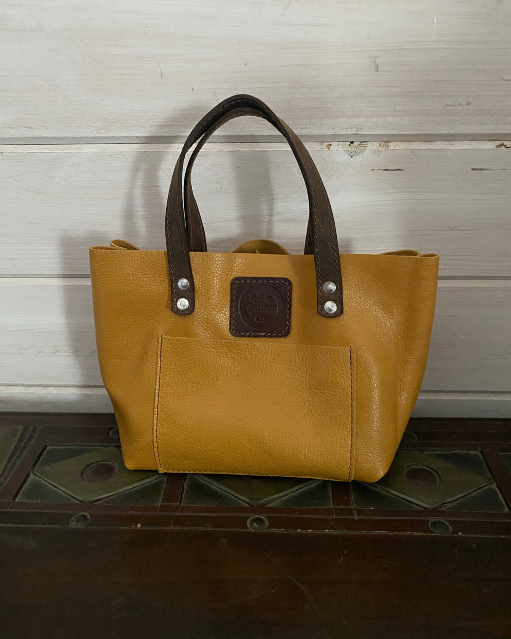 A tan leather tote bag with dark brown leather handles and a dark brown logo patch, shown hanging against a white wooden wall next to a dark wooden door.