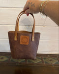 A brown embossed leather tote bag with tan handles and a circular logo patch on the front, held by a tattooed hand with bracelets against a white wood paneled wall.